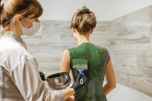 Young woman during mud treatment at spa, doctor applies green therapeutic mud on the patient's back at the treatment room photo