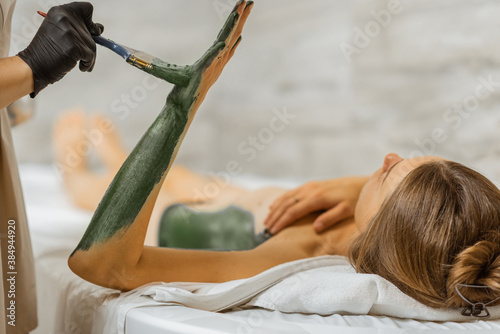 Young woman relaxing during a mud treatment at spa, doctor applies green therapeutic mud on the patient's body at the treatment room photo