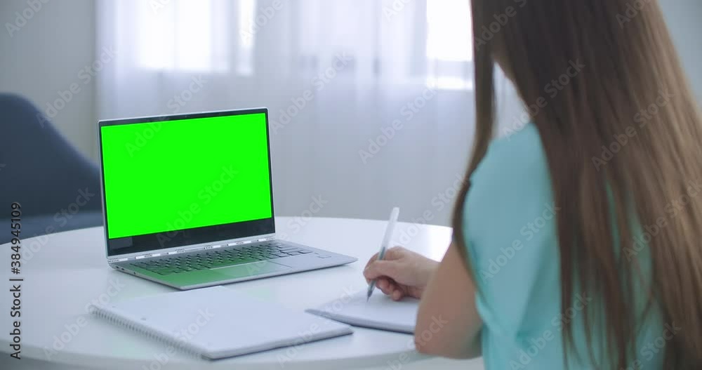Woman sits at desk in bedroom, she looks at laptop green screen and talks to someone over internet video communications, sometimes taking notes in notebook. Close-up Woman sits at desk in bedroom, she looks at laptop green screen and talks to someone over internet video communications, sometimes taking notes in notebook. Close-up