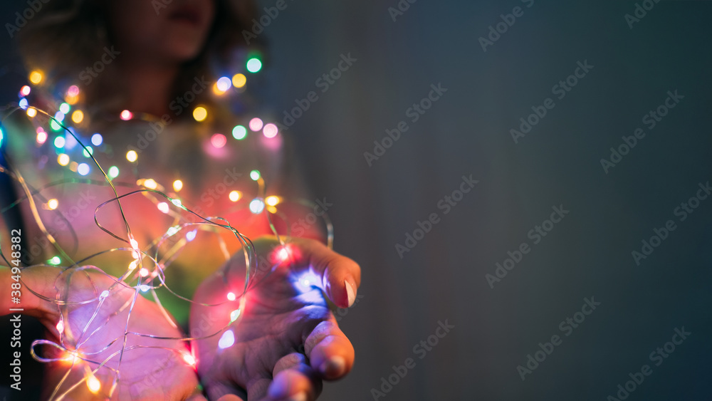 Christmas background. Party decoration. Winter holidays celebration. Colorful glowing bokeh lights in female hands with open palms on blur dark copy space.