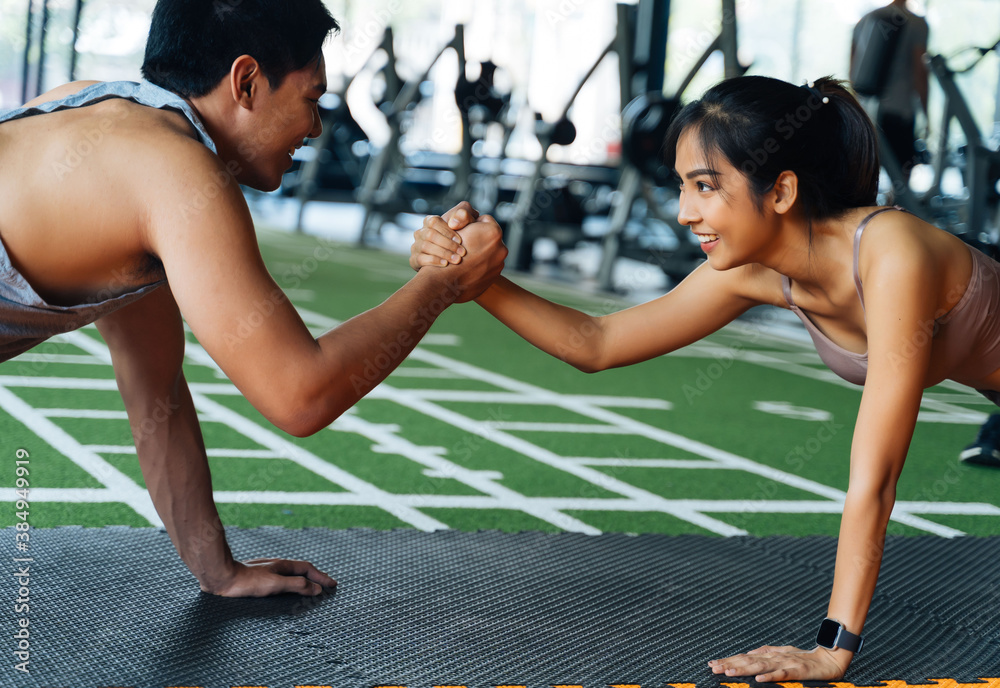 Healthy smiling couple of man and woman giving high five to each other while pushing up in the fitness gym. Asian sporty people working out together. Teamwork and achievement concept