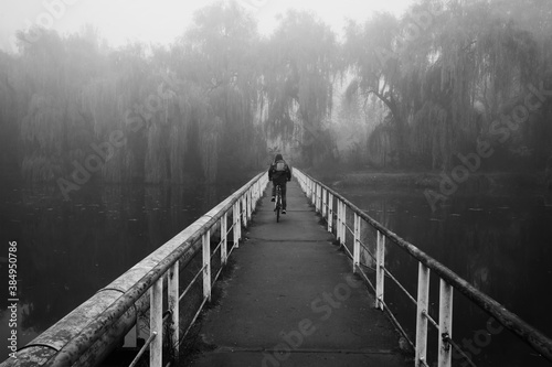 The man is cycling through the old rusty bridge over the lake in the early foggy morning