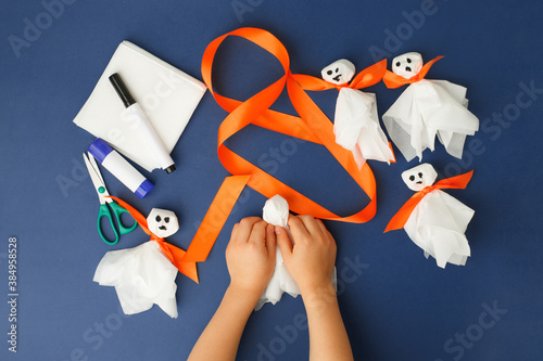 Boy makes Halloween toys ghost from white paper napkin. Creative DIY for kids on classic blue background. Home decor project party. Halloween crafts inspiration, recycle concept