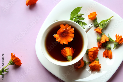 A Cup of tea with marigold flowers in a white dish with scattered flowers, purple background, top view, close - up-the concept of cooking and using herbal infusions