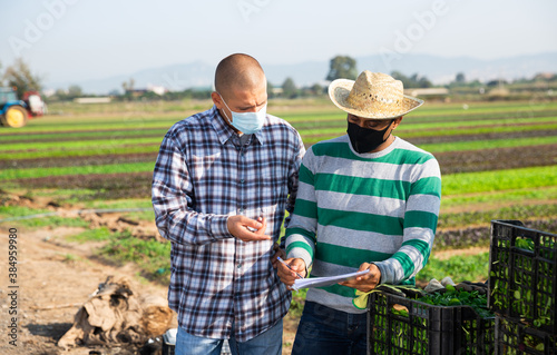 Hispanic farmer in protective face mask discussing work plan with his partner on farm field. Pandemic prevention and social distancing concept