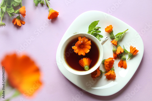 A Cup of tea with marigold flowers in a white dish with scattered flowers, purple background, top view, bokeh - the concept of cooking and using herbal infusions