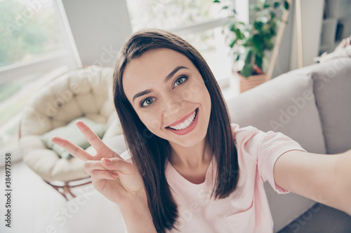 Photo portrait of overjoyed girl taking selfie showing v-sign with two fingers sitting on sofa indoors