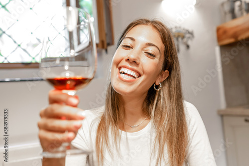 Cute young caucasian blonde woman toasting with red wine glass looking at the camera at home - New normal remote conference communication by online technology for the Coronavirus pandemic photo