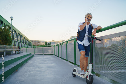 Portrait of mature man with electric scooter outdoors in city, using smartphone.