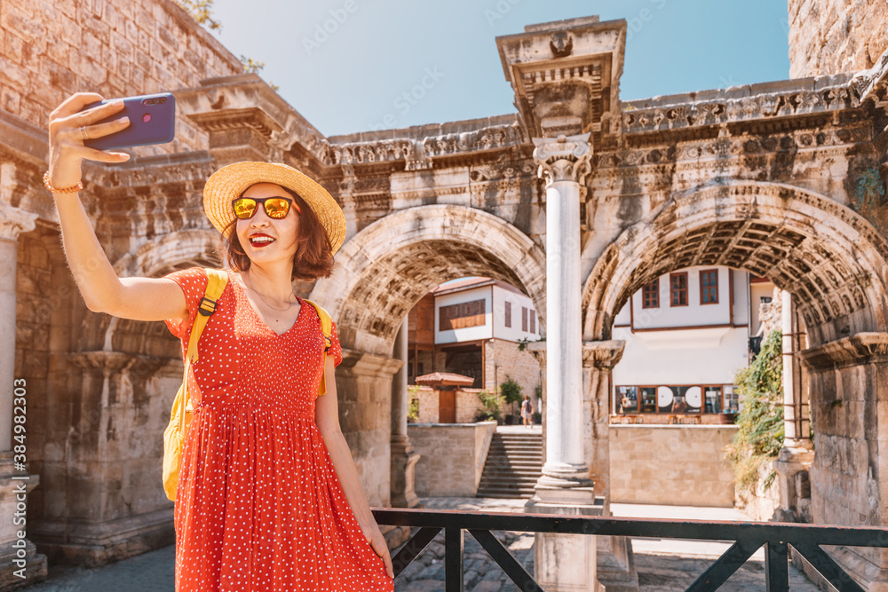 Happy female tourist traveller takes selfie photos against the backdrop of Hadrian's gate - a popular attraction in the old city of Antalya, Turkey