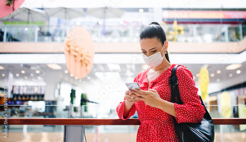 Woman with face mask standing with smartphone indoors in shopping center, coronavirus concept. photo