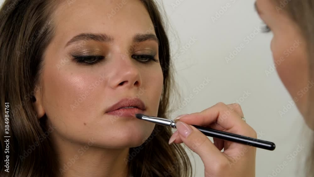 Beautiful well-groomed young woman applies stylish makeup to the model at beauty salon. Makeup artist making professional make-up and applying lipstick to the lips of the young woman Beautiful well-groomed young woman applies stylish makeup to the model at beauty salon. Makeup artist making professional make-up and applying lipstick to the lips of the young woman