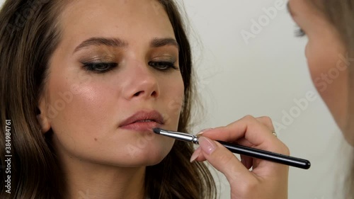 Beautiful well-groomed young woman applies stylish makeup to the model at beauty salon. Makeup artist making professional make-up and applying lipstick to the lips of the young woman