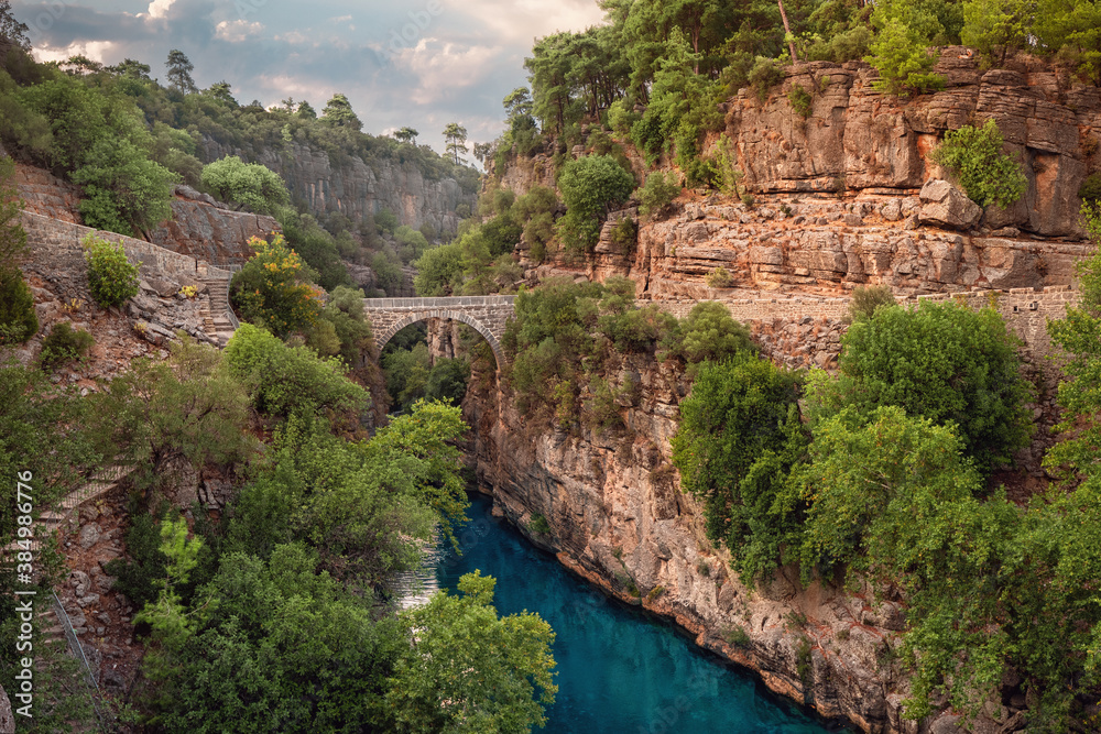 Fototapeta premium Ancient arch bridge over the Koprucay river gorge in Koprulu national Park in Turkey. Panoramic scenic view of the canyon and blue stormy mountain river
