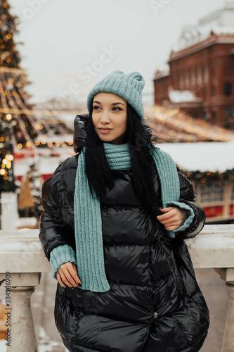 Outdoor photo of young beautiful girl with black hair in knitted blue hat and scarf, black jacket posing on street of city. Christmas, winter holidays concept. 