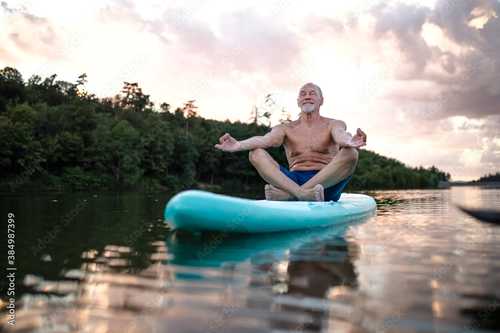 Senior man on paddleboard on lake in summer, doing yoga exercise.