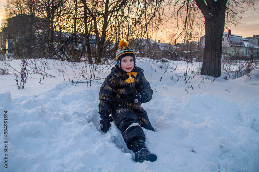 The kid in overalls is sitting in the snow and smiling. Portrait of a child in the snow.
