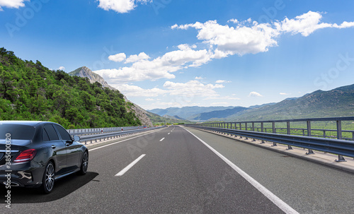 A Black car rushes along the road against the backdrop of a beautiful countryside landscape.