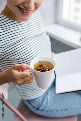 Cropped view of young woman holding cup of herbal tea and notebook at home