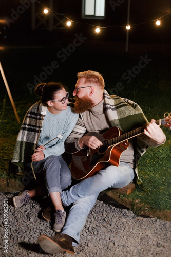 Young couple, caucasian woman and red haired bearded man, on a night picnic with a guitar and marshmello have a good time © vladdeep