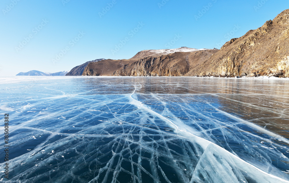 Frozen Baikal Lake in sunny cold February day. Beautiful winter landscape with transparent smooth ice near the rocky shore. Ice travel. Natural winter background