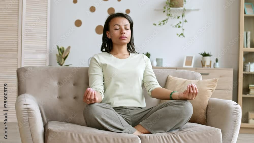 Lockdown of young pretty mixed-race woman wearing casual clothes sitting on couch in lotus position and meditating
