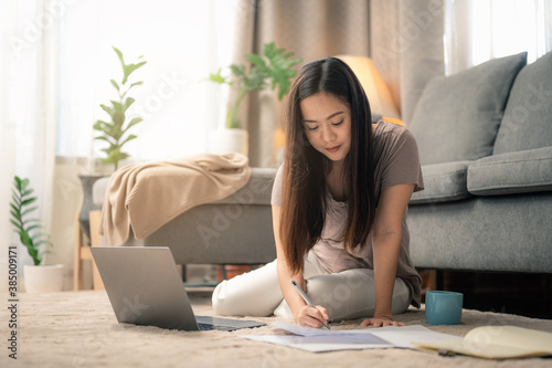 Portrait of smiling happy beautiful asian woman relaxing using technology of laptop computer while sitting on carpet.Young creative girl working and write on book in livingroom at home.