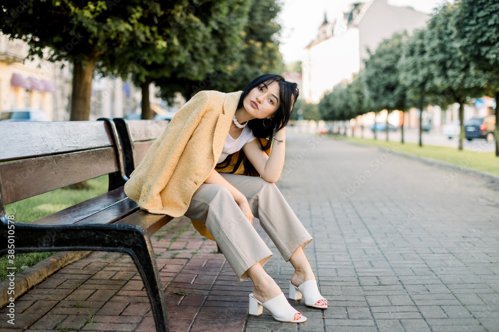 Beautiful brunette mixed raced sexy girl, wearing trendy yellow blazer and beige pants, posing to camera while sitting on the bench at city alley with green trees on the background Beautiful brunette mixed raced sexy girl, wearing trendy yellow blazer and beige pants, posing to camera while sitting on the bench at city alley with green trees on the background