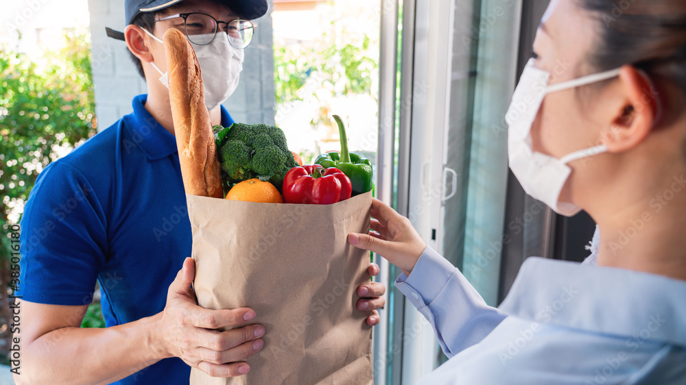 Asian deliver man wearing face mask in uniform handling bag of food and fruit give to woman costumer in front of the house, Express delivery service quarantine and shopping concept. Asian deliver man wearing face mask in uniform handling bag of food and fruit give to woman costumer in front of the house, Express delivery service quarantine and shopping concept.