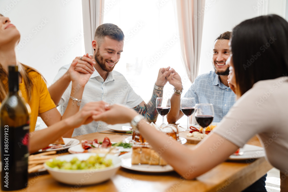 Image of happy multinational friends praying before lunch with holding hands together