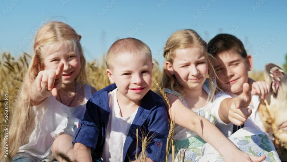 boy with down syndrome and healthy children show fingers at the camera and smiling, joyful friends sitting in a wheat field against the backdrop of a beautiful blue sky boy with down syndrome and healthy children show fingers at the camera and smiling, joyful friends sitting in a wheat field against the backdrop of a beautiful blue sky