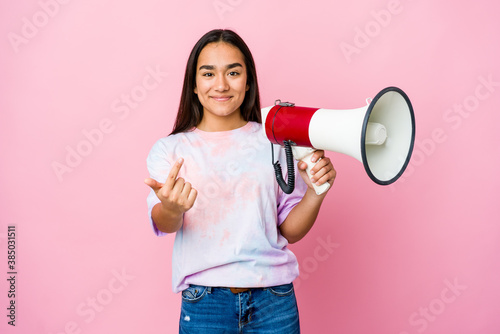 Young asian woman holding a megaphone isolated on pink background pointing with finger at you as if inviting come closer.