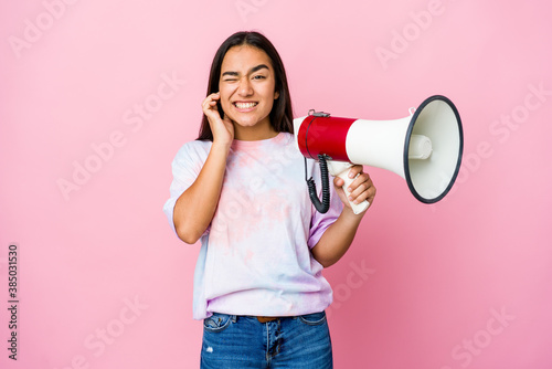 Young asian woman holding a megaphone isolated on pink background covering ears with hands.
