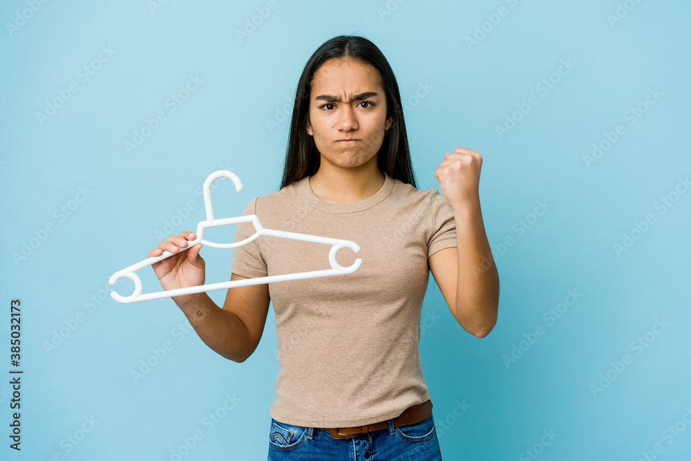 Young asian woman holding a hanger isolated on blue background showing fist to camera, aggressive facial expression. Young asian woman holding a hanger isolated on blue background showing fist to camera, aggressive facial expression.