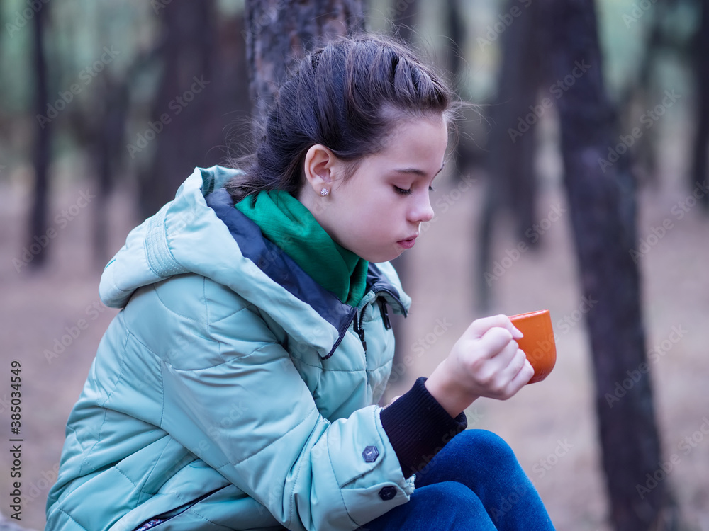 Plakat A teenage girl in the fall sits on a log and drinks hot tea from a mug. Rest in the forest.