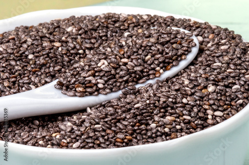 Chia seeds (Salvia hispanica) in white porcelain bowl and spoon, isolated on light blue background. Healthy food or superfoods and supplements concept. View from above. Flat lay.