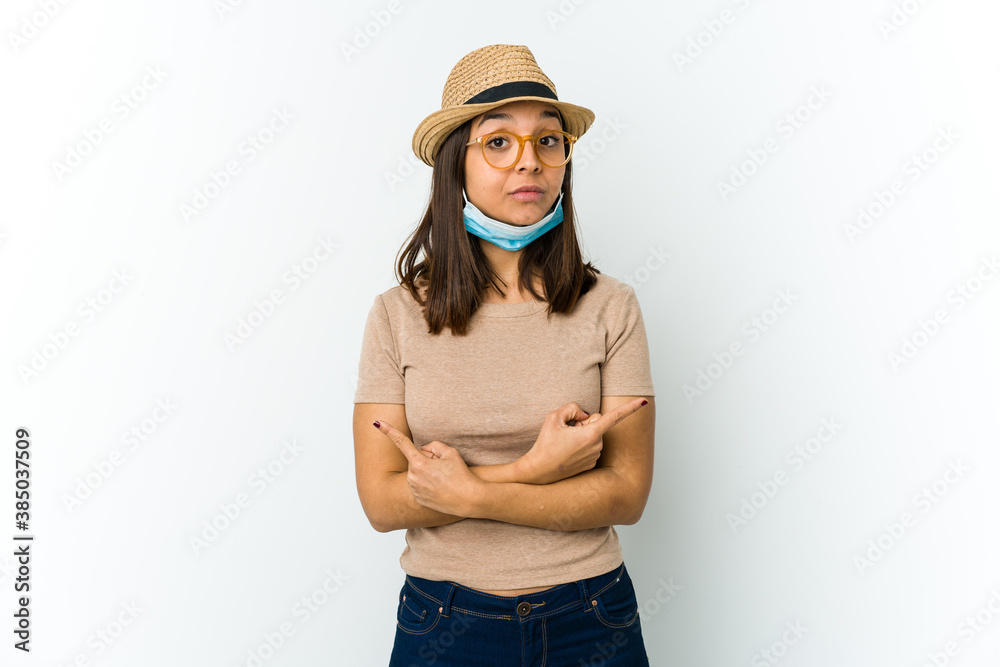Young latin woman wearing hat and mask to protect from covid isolated on white background points sideways, is trying to choose between two options. Young latin woman wearing hat and mask to protect from covid isolated on white background points sideways, is trying to choose between two options.
