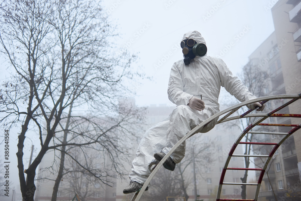 Man in gas mask and protective clothing monitoring city environment, holding tweezers. From below view of scientist sitting on ladder for kids on playground. Environmental disaster, smoky atmosphere. Man in gas mask and protective clothing monitoring city environment, holding tweezers. From below view of scientist sitting on ladder for kids on playground. Environmental disaster, smoky atmosphere.