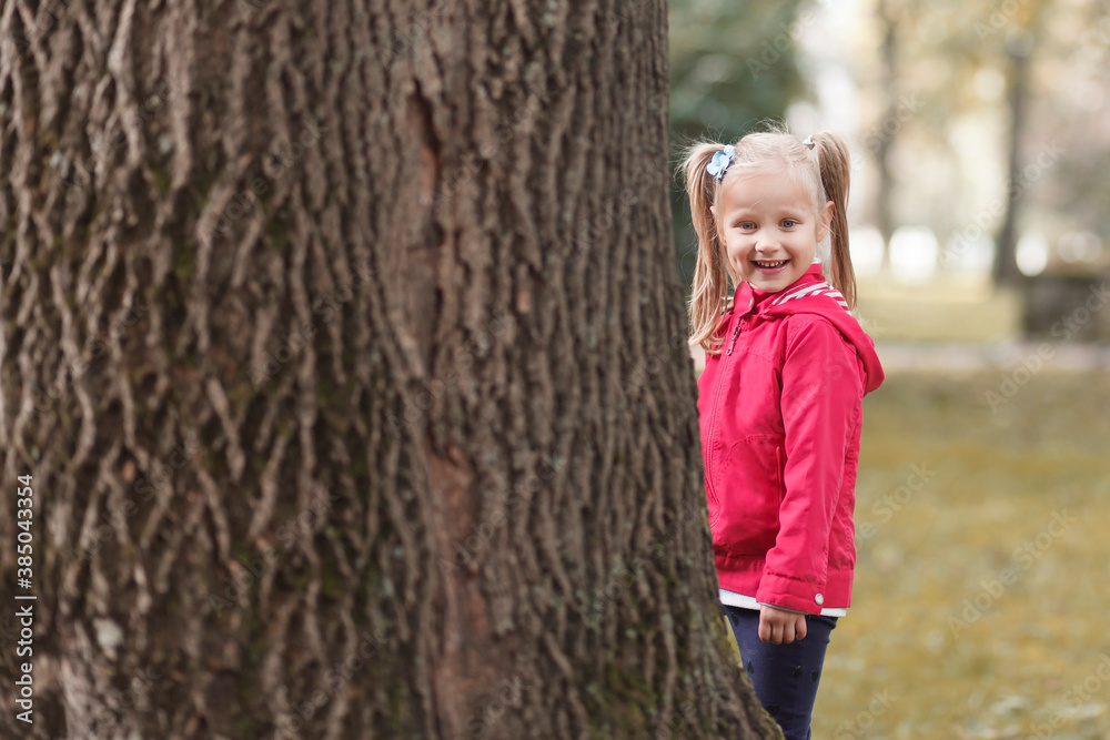 A little girl with two ponytails on her head stands near a wide tree trunk in the park