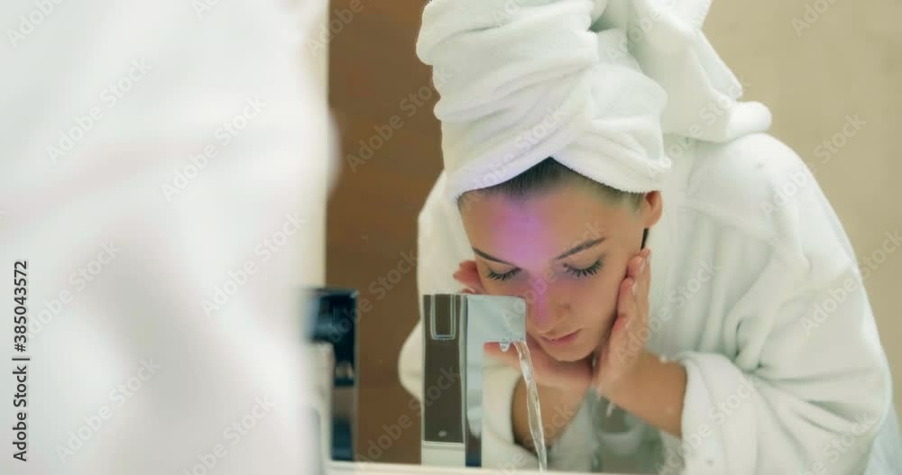 Young lady with white towel on head in bathrobe washes face looking at large mirror reflection in hotel bathroom closeup Young lady with white towel on head in bathrobe washes face looking at large mirror reflection in hotel bathroom closeup