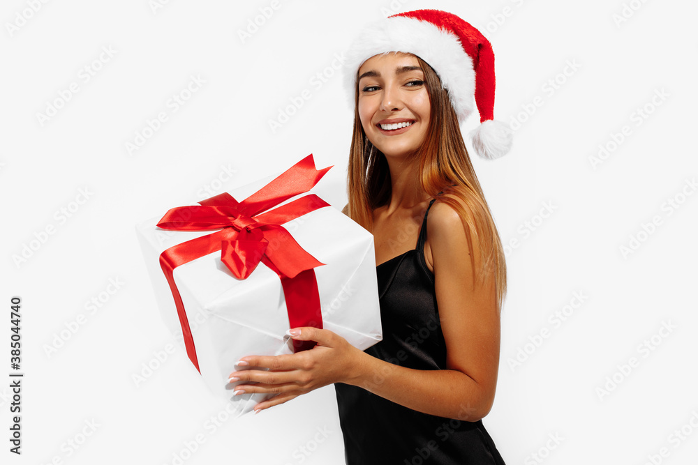 Happy beautiful young woman in a red Santa Claus hat and in a black elegant dress, with a gift on a white background, holiday concept, New Year, Christmas Happy beautiful young woman in a red Santa Claus hat and in a black elegant dress, with a gift on a white background, holiday concept, New Year, Christmas