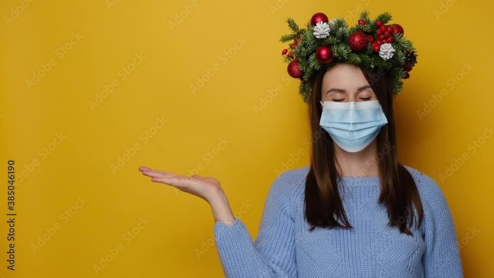 Young woman in traditional Christmas wreath wears respirator mask to protect herself from coronavirus, demonstrates empty copy space for promo text, isolated on yellow wall. Pandemic and New Year Young woman in traditional Christmas wreath wears respirator mask to protect herself from coronavirus, demonstrates empty copy space for promo text, isolated on yellow wall. Pandemic and New Year
