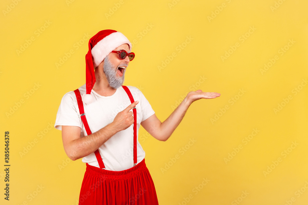 Positive elderly man in sunglasses and santa claus hat pointing finger showing freespace, advertisement area. Indoor studio shot isolated on yellow background Positive elderly man in sunglasses and santa claus hat pointing finger showing freespace, advertisement area. Indoor studio shot isolated on yellow background