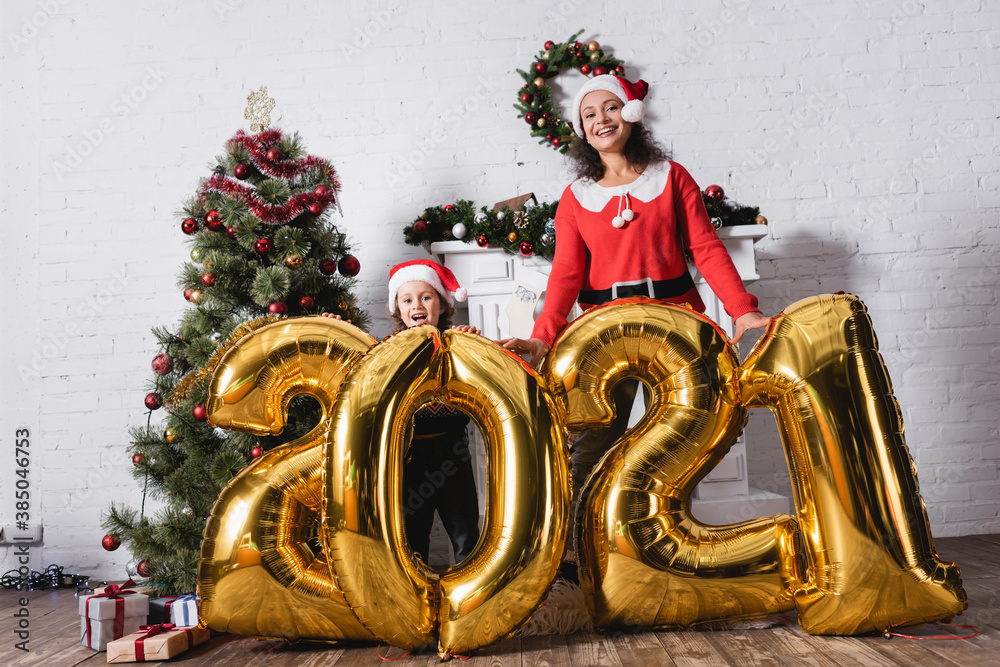 Daughter and mother in santa hats standing near balloons with numbers - obrazy, fototapety, plakaty 