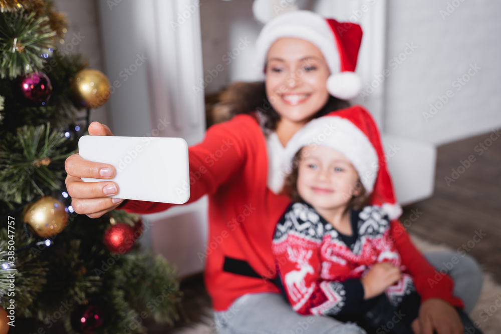 Selective focus of mother and daughter in santa hats taking selfie at home Selective focus of mother and daughter in santa hats taking selfie at home
