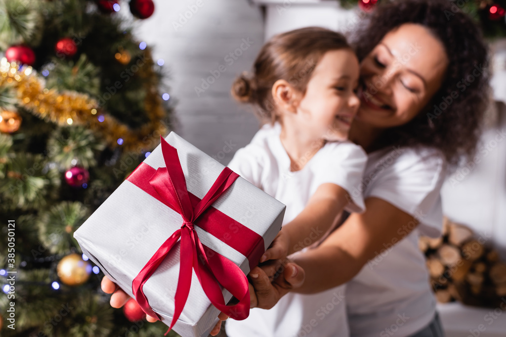 Selective focus of mother hugging daughter with gift box near decorated pine Selective focus of mother hugging daughter with gift box near decorated pine