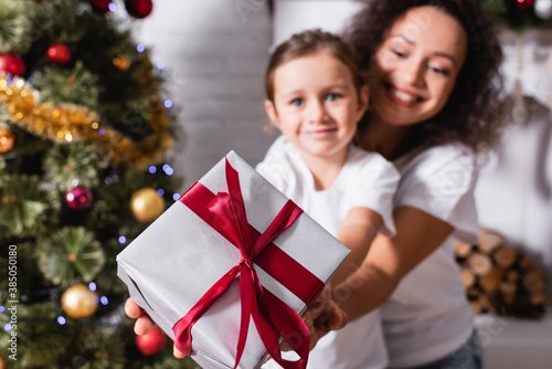 Selective focus mother hugging daughter with gift box near festive pine at home