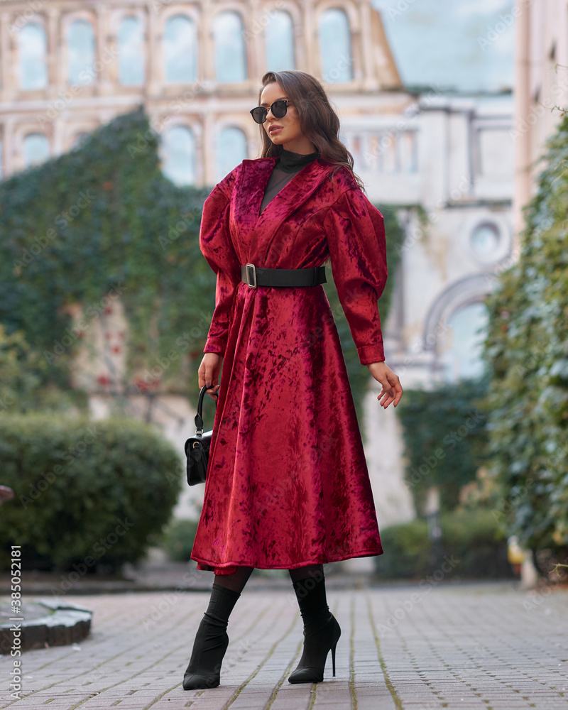 Stylish woman in red long coat with belt, black skirt, tights, sunglasses and high heels walking at city street. Brunette caucasian girl with long wavy hair outdoor portrait