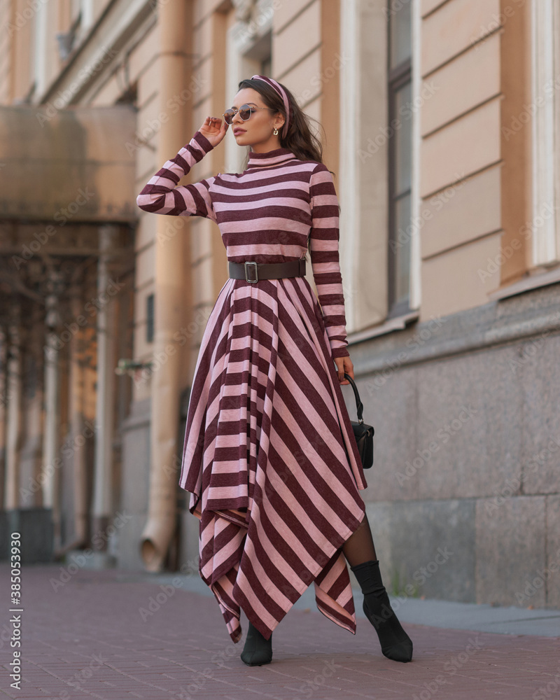 Elegant woman in long warm dress with pink and brown stripes walking at city street. Female model with long wavy brunette jair in sunglasses with black leather handbag