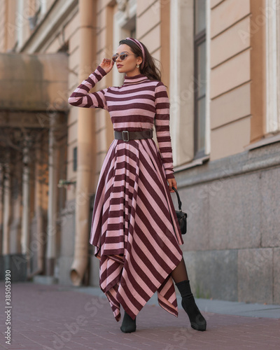 Elegant woman in long warm dress with pink and brown stripes walking at city street. Female model with long wavy brunette jair in sunglasses with black leather handbag